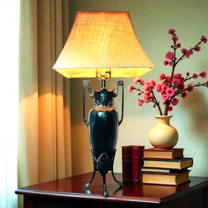 May include: A dark blue table lamp with a beige lampshade. The lamp has a decorative, vase-like base with ornate details. A vase with pink flowers and a stack of books are also on the table.