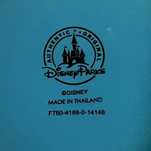 May include: A close-up of a teal surface with a black circular logo that says "AUTHENTIC ORIGINAL" around a castle silhouette. Below the logo is "Disney Parks" and the text "&copy;DISNEY MADE IN THAILAND F760-4188-0-14148".