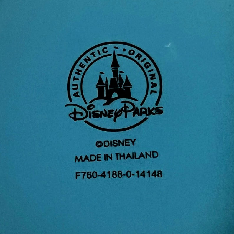May include: A close-up of a teal surface with a black circular logo that says "AUTHENTIC ORIGINAL" around a castle silhouette. Below the logo is "Disney Parks" and the text "&copy;DISNEY MADE IN THAILAND F760-4188-0-14148".