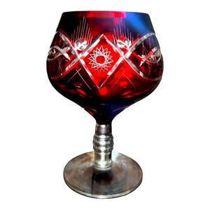 May include: A ruby red crystal brandy snifter with intricate etched designs. The glass features a wide bowl, a short stem, and a circular base. The etched patterns include diamond shapes and starbursts, adding a touch of elegance to the glassware.