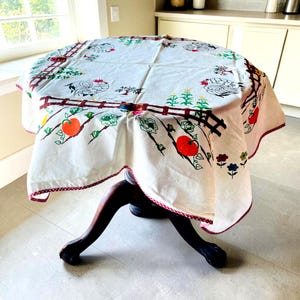 May include: A round white tablecloth featuring a printed pattern of roosters, brown fences, green trees, orange tomatoes, and small flowers. The edges are finished with a thin red and white checkered trim, draping over a dark wooden pedestal table.
