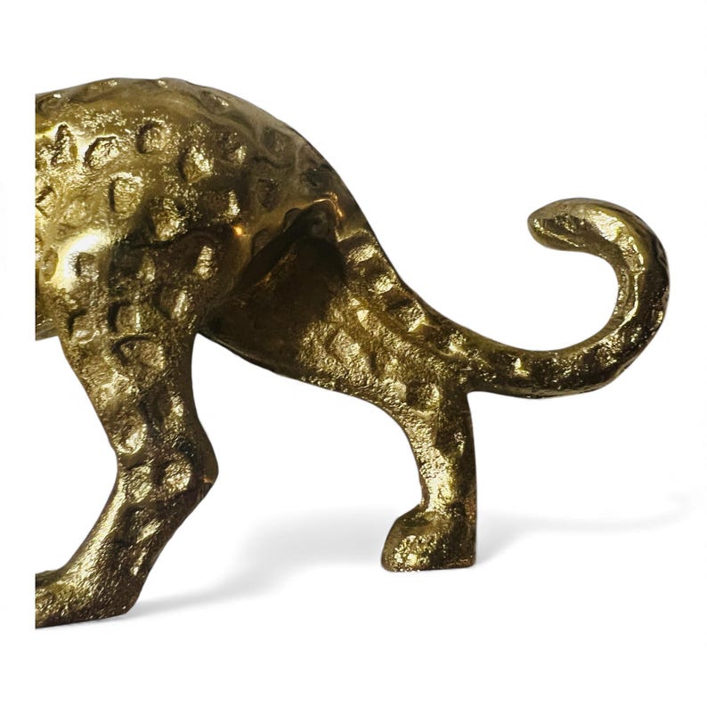 May include: A golden, textured metal sculpture of a cheetah, showcasing its hindquarters and curled tail. The sculpture has a hammered appearance, with visible spots and a polished finish. The cheetah's legs are firmly planted, suggesting a poised stance.