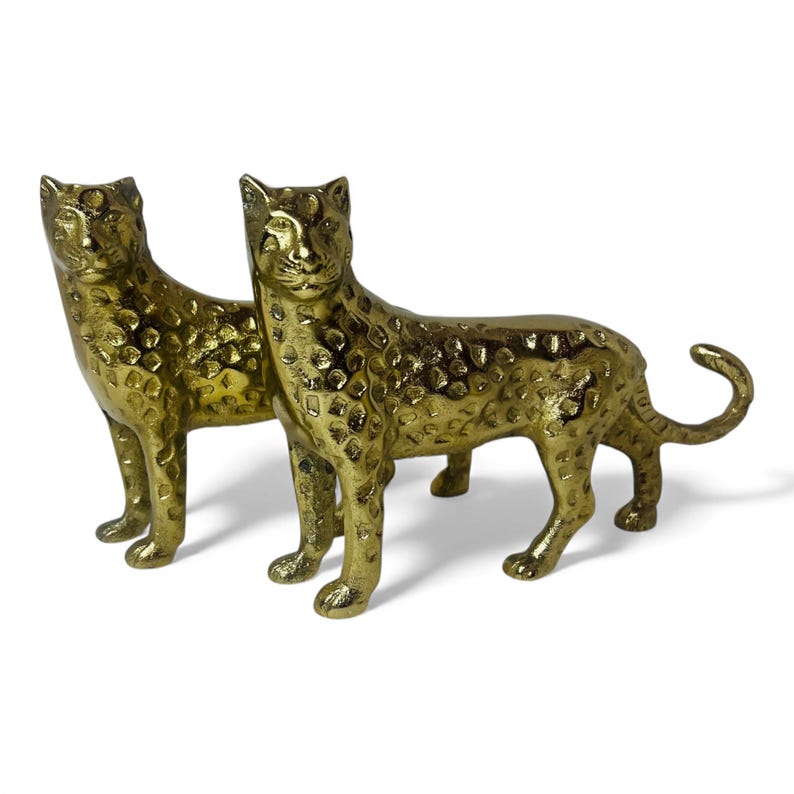 May include: Two gold-colored leopard figurines. The statues have a textured surface and are posed in a standing position. The leopards have a detailed design, including their spots and facial features. The tails are curled.