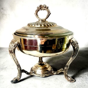 May include: Silver-plated chafing dish with a clear glass insert and ornate detailing. The lid features a decorative handle, and the dish rests on three curved legs. A classic serving piece for formal events.
