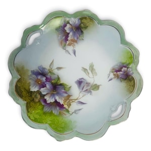 May include: A scalloped-edge decorative plate with a light green border and a floral design. The plate features hand-painted purple and white flowers with green leaves and stems. The background is a soft, light blue.