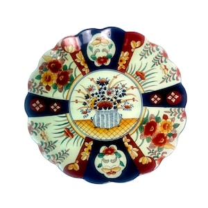 May include: A round ceramic plate with a scalloped edge featuring a central illustration of a blue and white patterned vase filled with flowers. The design includes alternating panels of deep blue and cream, decorated with red and yellow floral motifs.