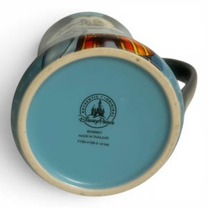 May include: A ceramic mug with a black handle and a light blue interior. The bottom of the mug is light blue with the "Authentic Disney Parks" logo and text. The mug features a cartoon character design with blue, red, and yellow accents.