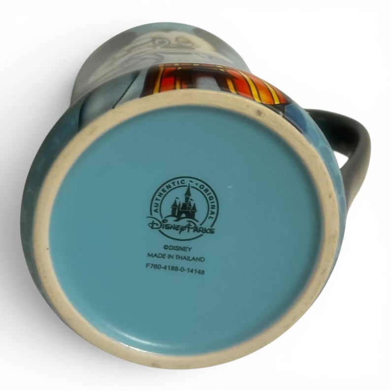 May include: A ceramic mug with a black handle and a light blue interior. The bottom of the mug is light blue with the "Authentic Disney Parks" logo and text. The mug features a cartoon character design with blue, red, and yellow accents.