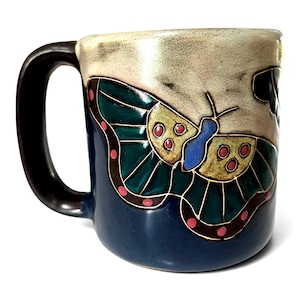 May include: A ceramic mug with a dark brown handle and a blue base. The mug features a detailed butterfly design with green, yellow, blue, and red accents. The top of the mug is a light beige color.