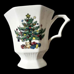 May include: A white ceramic mug with an octagonal shape and a faceted handle. The front features a colorful illustration of a decorated Christmas tree with a star on top, surrounded by ornaments, a teddy bear, and several wrapped gift boxes.