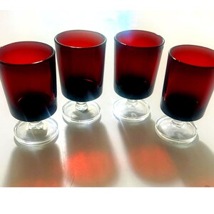 May include: A set of four matching drinking glasses, each featuring a deep red cylindrical bowl supported by a clear glass stem and a circular base. The glasses are arranged in a row, showcasing the contrast between the red glass and clear base.