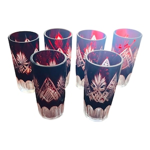 May include: Six ruby red and clear cut crystal drinking glasses. The glasses have a tall, cylindrical shape with intricate geometric and floral patterns. The interior of the glasses is a deep red, contrasting with the clear, cut exterior.