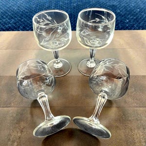 May include: Four clear glass wine glasses with etched floral designs. Two glasses are upright, while the other two are lying on their sides. The glasses have a classic stem and a round base. The background is a dark brown surface.