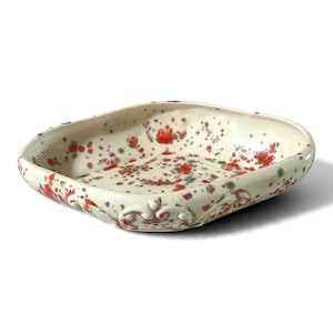 May include: A shallow, square ceramic dish with rounded corners and a cream-colored base. The surface is decorated with a speckled pattern of red and green paint splatters. The exterior rim features a raised, molded scrollwork detail.