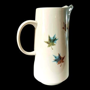 May include: Cream-colored ceramic pitcher with a curved handle and spout. The pitcher is decorated with three stylized leaf designs in shades of green, blue, and brown. The pitcher is a vintage style.