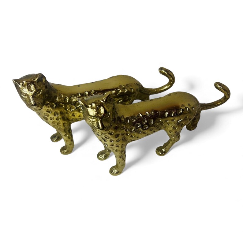 May include: Two gold-colored leopard figurines. The figurines are side-by-side, with textured surfaces and spotted patterns. The leopards are in a walking pose, with their tails curled upwards. The background is white.
