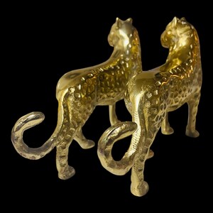 May include: Two golden cheetah figurines with textured surfaces stand side-by-side against a black background. The cheetahs are posed with their tails curled, showcasing their elegant form. These decorative sculptures add a touch of luxury.