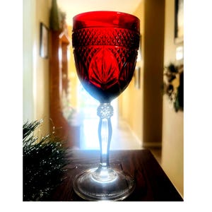 May include: A ruby red wine glass with a decorative cut glass design. The bowl features a diamond pattern and a central fan design. The stem has a crystal-like embellishment. The glass sits on a clear, round base. The glass is on a dark wooden surface.