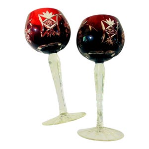 May include: Two wine glasses featuring deep red bowls with intricate clear cut-glass star and floral patterns. Each glass has a long, slender, pale yellow stem with a textured surface and a wide, clear circular base for stability and balance.