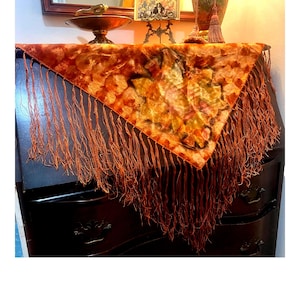 May include: A decorative, triangular scarf with a floral pattern in shades of orange, yellow, and brown, draped over a dark wooden dresser. The scarf has long, orange fringe along the edges, adding a vintage touch.