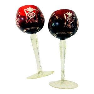 May include: Two wine glasses featuring deep red bowls with intricate cut-glass star and floral patterns. Each glass has a pale yellow, textured stem and a wide, clear circular base. The design showcases a contrast between the dark red upper section and the light-colored stem, creating a distinct visual separation in the glassware.