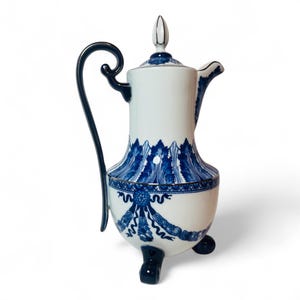 May include: A white and blue porcelain coffee pot with a curved handle and a decorative blue floral pattern. The pot has a lid with a small finial and rests on three small feet. The design features a blue and white color scheme.
