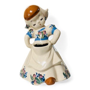 May include: A ceramic figurine of a girl in a white dress with blue and pink floral accents. She is holding her hands in her apron, which has a small opening for a flower pot.