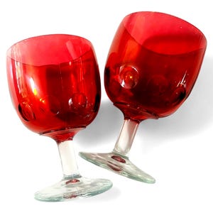 May include: Two red wine glasses with clear stems and bases are angled towards the viewer. The red bowls have a rounded shape with a small, circular indentation. The glasses are set against a white background.
