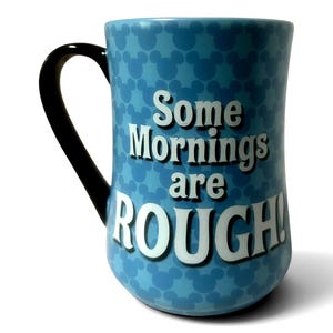 May include: A light blue ceramic mug with a black handle. The mug features a pattern of Mickey Mouse silhouettes and the text "Some Mornings are ROUGH!" in white block letters.