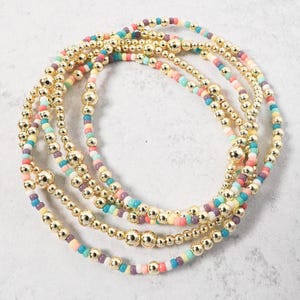 May include: Gold beaded bracelets with colorful beads in pastel shades of pink, yellow, blue, and purple. The bracelets have a mix of small and large gold beads. The bracelets are arranged in a circular pattern.