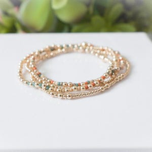 May include: A set of gold beaded bracelets with small, round beads. Some bracelets have accent beads in orange and teal. The bracelets are stacked on a white surface, with a blurred green background.