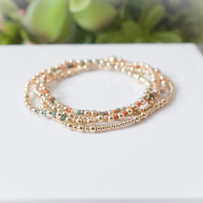 May include: A set of gold beaded bracelets with small, round beads. Some bracelets have accent beads in orange and teal. The bracelets are stacked on a white surface, with a blurred green background.