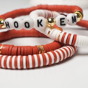 May include: Three handmade bracelets in shades of red and white. One bracelet spells out "HOOKEM" with black letters on white beads. The other bracelets have gold accents and a mix of solid and striped designs.