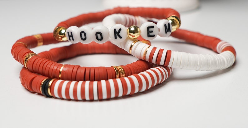 May include: Three handmade bracelets in shades of red and white. One bracelet spells out "HOOKEM" with black letters on white beads. The other bracelets have gold accents and a mix of solid and striped designs.