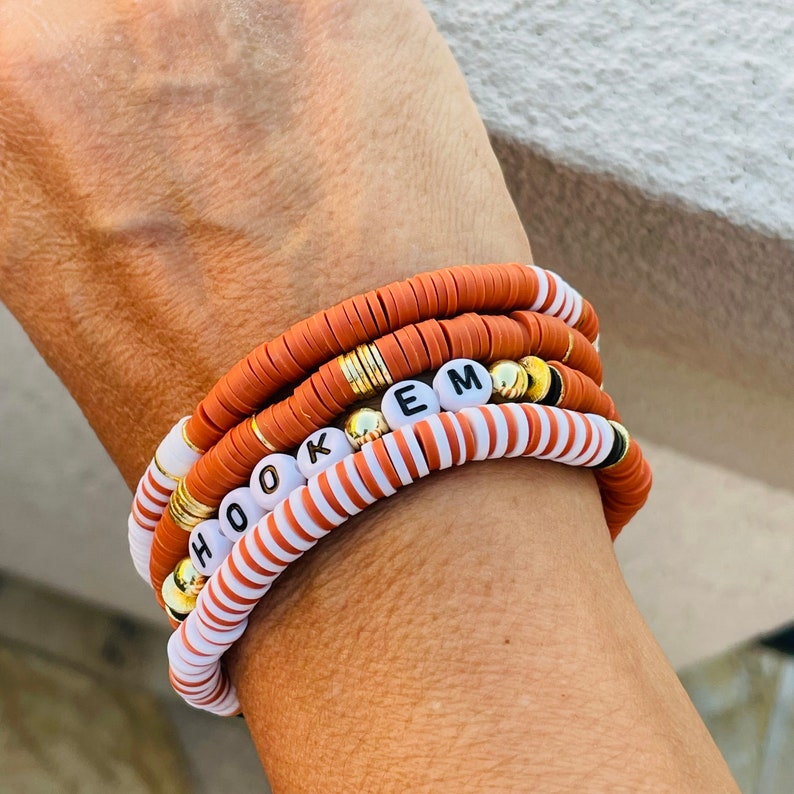 May include: Stack of four bracelets in shades of orange, white, and gold. One bracelet spells out the word "HOOKEM" with white letter beads. The bracelets are made of flat, disc-shaped beads and gold accents.