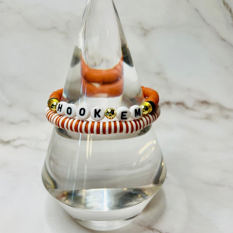 May include: A stack of two bracelets. The top bracelet is a burnt orange color with the word "HOOKEM" spelled out in white letter beads. The bottom bracelet has alternating white and burnt orange stripes. Gold-colored beads are interspersed.