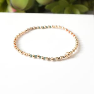 May include: Gold beaded bracelet with small accent beads in shades of orange, teal, and gray. The bracelet is made of small, round gold beads and has a single larger gold bead. The bracelet is on a white surface.