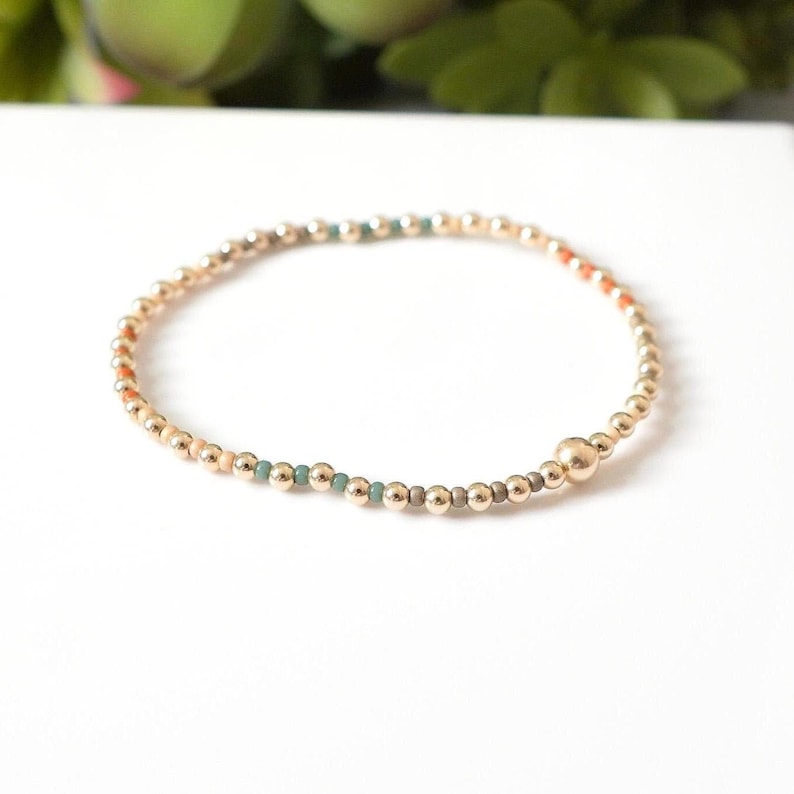 May include: Gold beaded bracelet with small accent beads in shades of orange, teal, and gray. The bracelet is made of small, round gold beads and has a single larger gold bead. The bracelet is on a white surface.