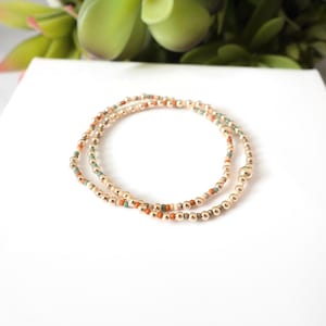 May include: Two gold beaded bracelets with small, colorful beads in orange, teal, and cream. The bracelets are displayed on a white surface, with green foliage in the background.