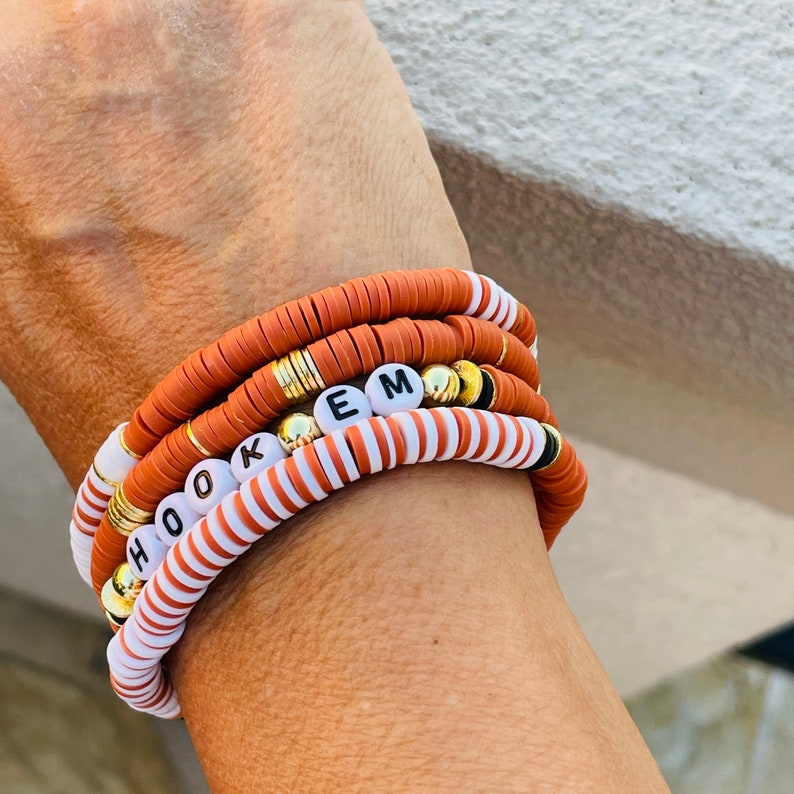 May include: Stack of four bracelets with a mix of terracotta, white, and gold beads. One bracelet spells out "HOOK EM" with white letter beads. The bracelets are worn on a wrist.