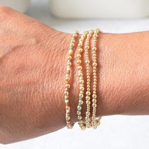 May include: A collection of gold beaded bracelets. One bracelet features a mix of orange, teal, and cream-colored beads. The other bracelets are made of solid gold beads. The bracelets are stacked on a wrist.