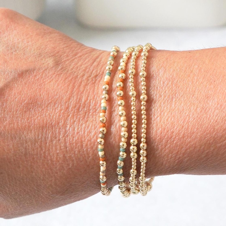 May include: A collection of gold beaded bracelets. One bracelet features a mix of orange, teal, and cream-colored beads. The other bracelets are made of solid gold beads. The bracelets are stacked on a wrist.