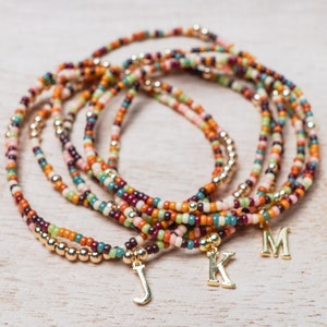 Op de afbeelding: Meerdere kralenarmbanden in verschillende kleuren, waaronder oranje, groen en bordeaux, zijn op elkaar gestapeld. Elke armband heeft een goudkleurige bedel met een letter. De armbanden liggen op een lichtgekleurd houten oppervlak.