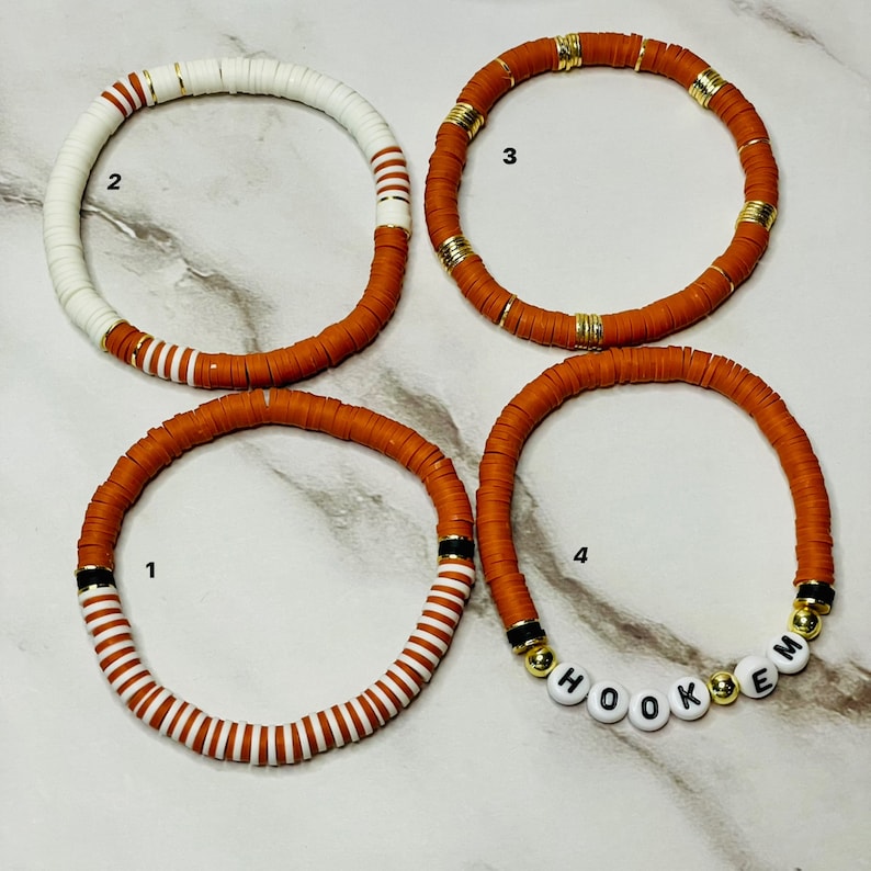 May include: Four handmade bracelets. The bracelets are made of small, flat, round beads in shades of white, orange, and gold. One bracelet spells out the word "HOOKEM" with white letter beads. The bracelets are a stylish accessory.