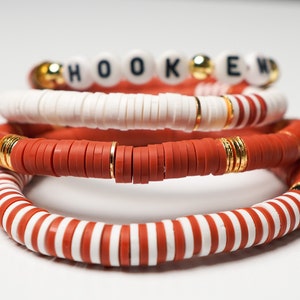 May include: Stack of four beaded bracelets. The top bracelet spells out "HOOKED" with white letter beads. Other bracelets are red, white, and gold, with a striped pattern on the bottom bracelet.