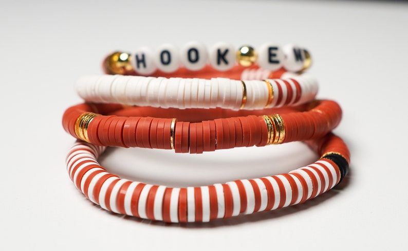 May include: Stack of four beaded bracelets. The top bracelet spells out "HOOKED" with white letter beads. Other bracelets are red, white, and gold, with a striped pattern on the bottom bracelet.