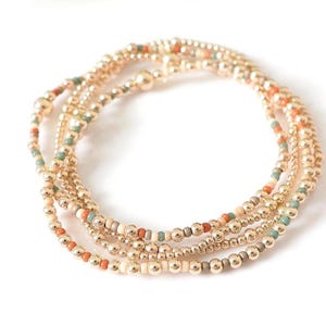 May include: Gold beaded bracelets with accents of orange, teal, and cream-colored beads. The bracelets are arranged in a circular pattern against a white background. The beads are small and evenly spaced.