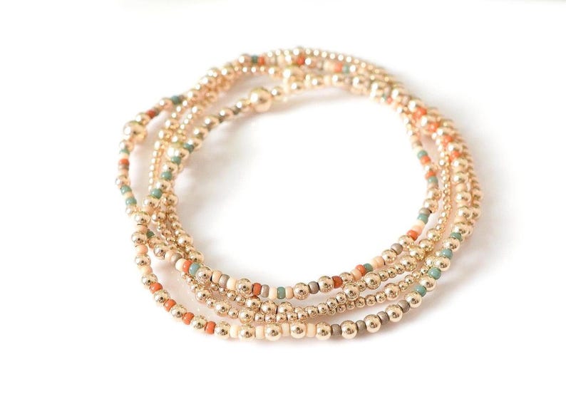 May include: Gold beaded bracelets with accents of orange, teal, and cream-colored beads. The bracelets are arranged in a circular pattern against a white background. The beads are small and evenly spaced.