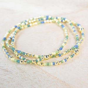 May include: Three beaded bracelets with a mix of small, colorful beads in yellow, green, and blue, interspersed with larger, gold-colored beads. The bracelets are arranged in a layered design on a light-colored wooden surface.