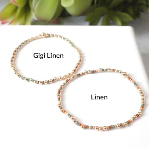 May include: Two beaded bracelets. One bracelet is gold with the text "Gigi Linen". The other bracelet is gold with the text "Linen". The bracelets have a mix of gold, beige, teal, and orange beads.